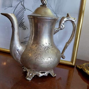 Può includere: Una caffettiera antica color argento con beccuccio curvo e manico ornato. La caffettiera presenta disegni floreali incisi e poggia su tre piedini decorativi. Uno schizzo incorniciato è sullo sfondo.