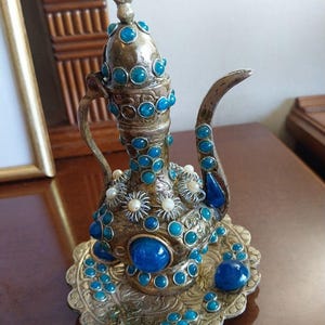 Puede incluir: Una tetera plateada ornamentada con detalles de piedras preciosas turquesas y azules. La tetera presenta un remate de media luna y está decorada con detalles florales y perlas. Se encuentra sobre un platillo a juego, con intrincados patrones.