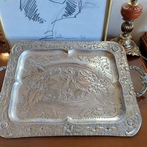 Puede incluir: Una bandeja rectangular de color plateado con intrincados diseños florales y de hojas. La bandeja tiene dos asas con adornos decorativos. Los bordes de la bandeja también están decorados con diseños florales. La bandeja es probablemente de metal.