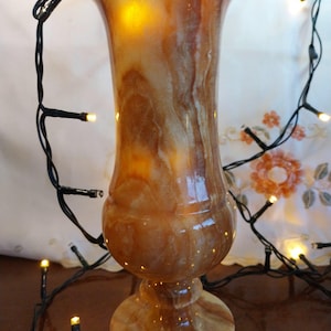 May include: A tall, polished stone vase featuring swirling patterns of amber, brown, and cream colors. The vase has a wide, flared rim, a rounded body, and a circular pedestal base. It is illuminated from within, casting a warm, glowing light.