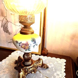Puede incluir: Una lámpara de mesa con una pantalla de cristal acanalado brillante sobre una base de cerámica blanca decorada con un patrón de rosas. Incluye un cuello de metal dorado con diseño de greca y una base de trípode con patas de garra.