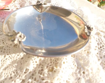 May include: A polished silver-toned metal dish with a smooth, reflective circular basin. The rim of the dish features three small, detailed floral accents spaced evenly around the edge, providing a decorative touch to the simple, shiny surface.