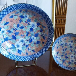 May include: Two round ceramic plates featuring a central design of blue and white flowers with green leaves set against a peach-colored background. The rims are decorated with a blue and white geometric grid pattern, and the plates rest on stands.