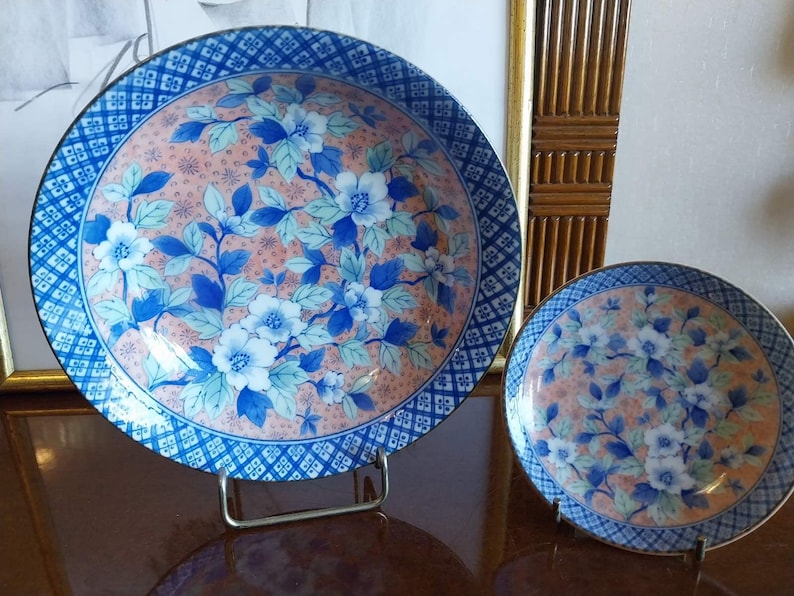 May include: Two round ceramic plates featuring a central design of blue and white flowers with green leaves set against a peach-colored background. The rims are decorated with a blue and white geometric grid pattern, and the plates rest on stands.