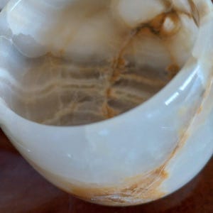 May include: A small, rounded vessel carved from translucent white onyx stone. The material features natural, swirling bands of cream and golden-brown mineral deposits throughout its smooth, polished surface, creating a unique, organic pattern.