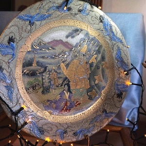 Könnte beinhalten: Dekorative Keramikplatte mit detailliertem asiatisch inspiriertem Design. Die Platte zeigt eine zentrale Szene mit Figuren, Bergen und einer Pagode, umgeben von einem Rand mit blauen Vögeln. Goldakzente verstärken das Design.