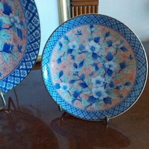 May include: A round ceramic plate featuring a central design of blue and white flowers with green leaves set against a soft peach-colored background. The rim is decorated with a blue and white geometric diamond pattern and a thin metallic gold edge.