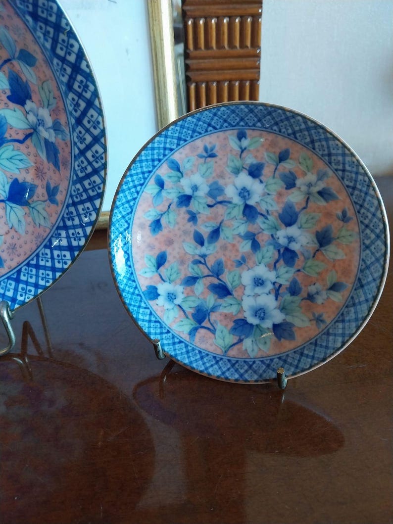 May include: A round ceramic plate featuring a central design of blue and white flowers with green leaves set against a soft peach-colored background. The rim is decorated with a blue and white geometric diamond pattern and a thin metallic gold edge.