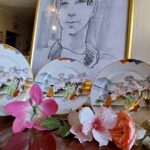 Puede incluir: Tres platos decorativos de porcelana con un diseño de paisaje japonés que presenta figuras con vestimenta tradicional, montañas y edificios. Los platos tienen un borde dorado y se exhiben con flores artificiales rosas y naranjas.