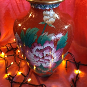 Pode incluir: Um vaso cloisonné vermelho com uma borda dourada e um padrão floral decorativo com grandes peónias cor-de-rosa e brancas, folhas verdes e um botão de flor branca mais pequeno. A superfície é adornada com um padrão de rede de fio de ouro.