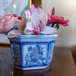Pode incluir: Um pequeno vaso hexagonal de cerâmica com um design floral azul e branco. O vaso contém um buquê de flores rosa e vermelhas, incluindo rosas e lírios. O vaso está sobre uma superfície de madeira.
