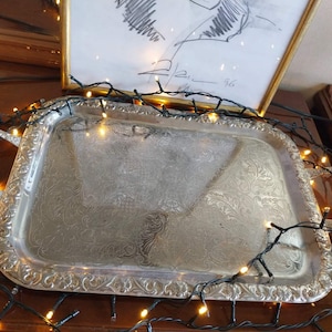 May include: A rectangular silver-toned serving tray featuring ornate embossed floral patterns along the border and handles. The tray is surrounded by a string of small, glowing warm white LED lights that illuminate the metallic surface.