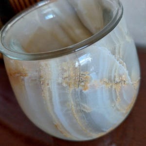 May include: A rounded vessel crafted from polished banded stone, featuring a translucent appearance with swirling layers of cream, white, and amber-colored veins. The container has a smooth, curved exterior and a wide, open circular rim.