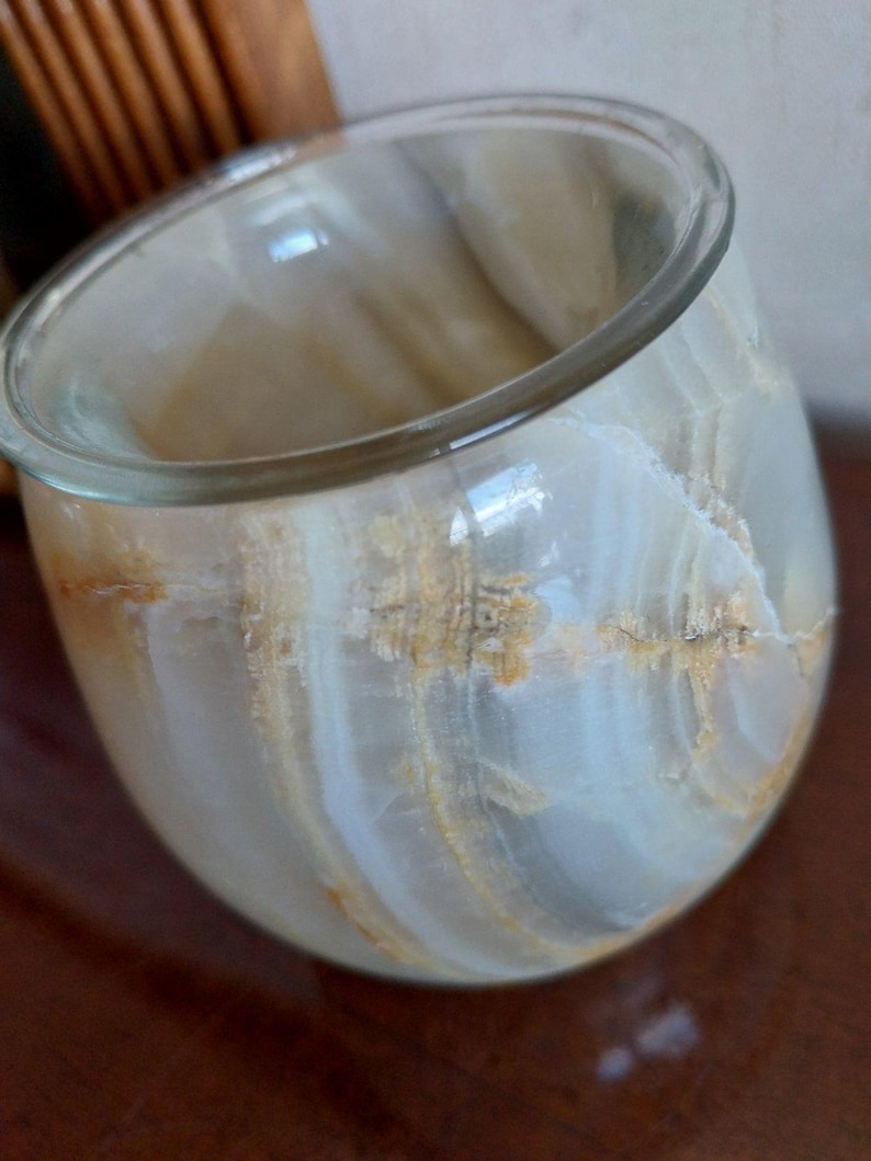 May include: A rounded vessel crafted from polished banded stone, featuring a translucent appearance with swirling layers of cream, white, and amber-colored veins. The container has a smooth, curved exterior and a wide, open circular rim.
