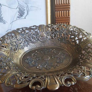 May include: An oval brass bowl featuring an intricate openwork filigree border with scrolling patterns and a central base decorated with embossed floral motifs and a cross-hatched texture. The metal has a dark, aged patina finish throughout.