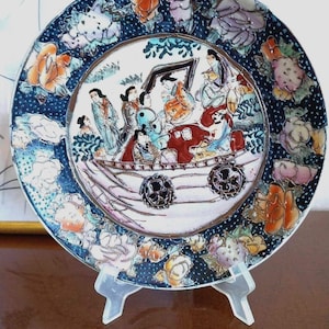 May include: A round ceramic decorative plate featuring a central illustration of multiple figures in a boat. The border is dark blue with a white dotted pattern and large, colourful floral motifs in shades of orange, pink, and white.