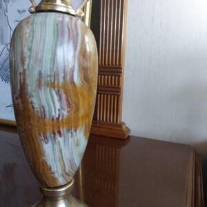 May include: A decorative vase with a gold-colored top and base. The body of the vase features a striped pattern in shades of brown, white, and green. The vase has two decorative handles and is displayed on a dark brown surface.