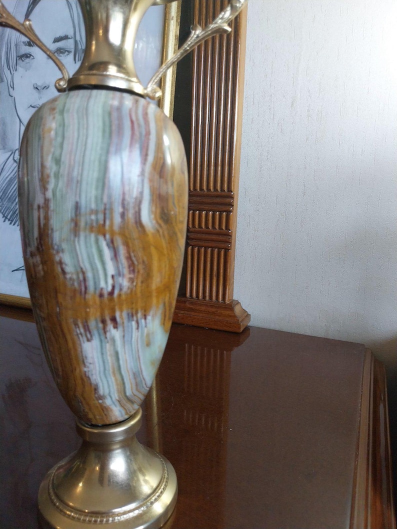 May include: A decorative vase with a gold-colored top and base. The body of the vase features a striped pattern in shades of brown, white, and green. The vase has two decorative handles and is displayed on a dark brown surface.