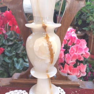 Può includere: Un vaso in pietra alto e cilindrico realizzato in onice lucido di colore bianco e crema. Il vaso presenta un bordo svasato, una sezione centrale bulbosa e una base circolare, il tutto accentuato da evidenti venature minerali marroni.