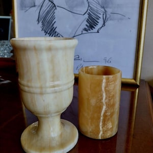 May include: A polished light beige marble chalice with a flared rim and a sturdy pedestal base, positioned next to a cylindrical amber-colored stone tumbler. Both vessels feature natural veining and a smooth, glossy finish throughout their surfaces.
