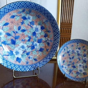 May include: Two round ceramic plates featuring a peach-colored center with blue and white floral patterns. The rims have a dark blue and white geometric grid design. Each plate is displayed on a metal stand, showcasing the detailed botanical art.