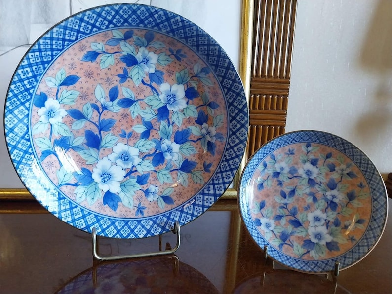 May include: Two round ceramic plates featuring a peach-colored center with blue and white floral patterns. The rims have a dark blue and white geometric grid design. Each plate is displayed on a metal stand, showcasing the detailed botanical art.