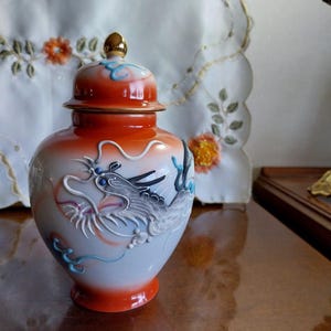 May include: A ceramic jar with a lid, featuring a dragon design. The jar has a white base with an orange gradient, and the lid is orange with a gold finial. The dragon is detailed in white and grey. Likely used for storage or display.