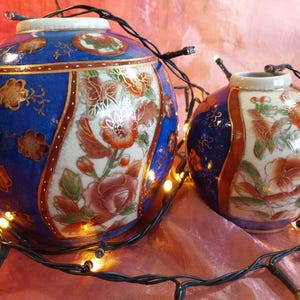 Puede incluir: Dos jarrones esféricos de cerámica con una vibrante paleta de colores azul y blanco con intrincados patrones florales en naranja, oro y verde. Los jarrones están decorados con paneles con bordes dorados y rodeados por una cadena de luces cálidas.