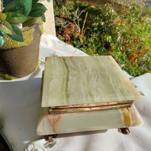 May include: A rectangular light green onyx stone box with natural white and orange veining. The box features a hinged lid with a decorative gold-toned metal trim and rests on four small, ornate gold-toned metal feet at the base corners.
