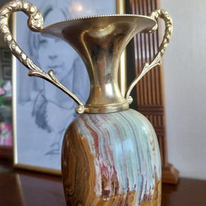 May include: A decorative vase with a gold-colored metal top and handles. The vase's body is made of stone with a mix of brown, green, and red hues. The handles curve upwards, adding a classic touch to the design.