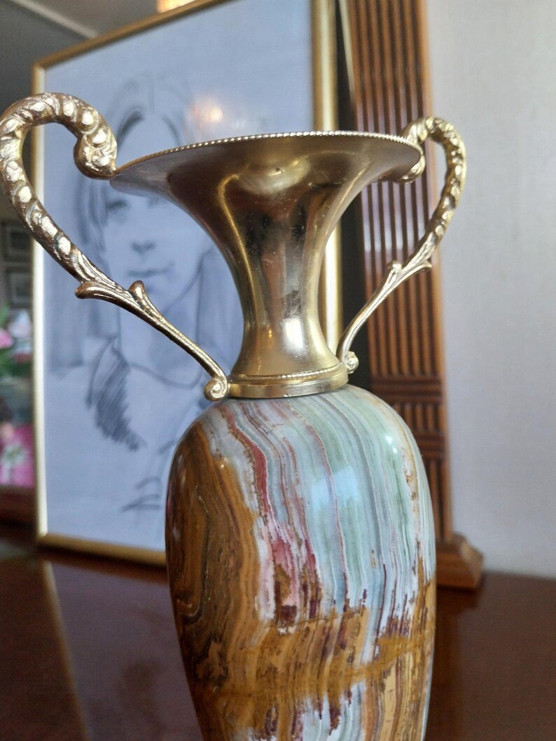 May include: A decorative vase with a gold-colored metal top and handles. The vase's body is made of stone with a mix of brown, green, and red hues. The handles curve upwards, adding a classic touch to the design.
