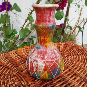May include: A hand-painted ceramic vase with a vibrant, abstract design. The vase features a red base with splashes of yellow, blue, and white. A white string net pattern decorates the lower portion. The vase is approximately 25 cm tall.