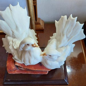 May include: A white porcelain figurine featuring two doves with detailed feathers and golden beaks, perched together on a textured, reddish-brown base. The birds are positioned facing each other with their tails fanned out in an upright display.