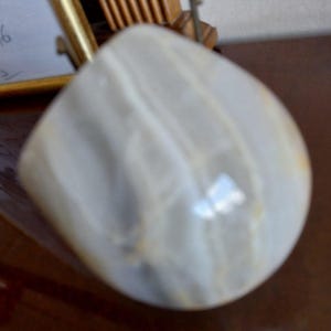 May include: A smooth, polished stone paperweight featuring a rounded, asymmetrical shape with a translucent, milky white appearance and subtle, soft gray banding patterns running throughout the surface of the mineral specimen.