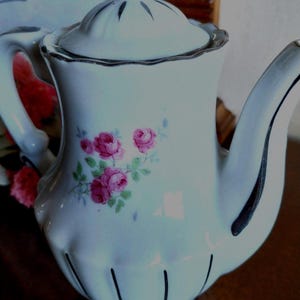 Peut inclure: Une théière en porcelaine blanche ornée d'un motif floral de roses roses délicates avec des feuilles vertes. La théière présente une base cannelée, un couvercle assorti avec un bouton, et des bordures argentées sur l'anse, le bec et le couvercle.