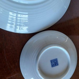 May include: Two white ceramic plates with a subtle ribbed texture on the underside. The smaller plate features a square blue stamp with Japanese characters centered on the base. The surfaces are smooth, glossy, and reflective, showing light.