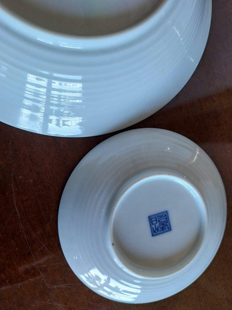 May include: Two white ceramic plates with a subtle ribbed texture on the underside. The smaller plate features a square blue stamp with Japanese characters centered on the base. The surfaces are smooth, glossy, and reflective, showing light.