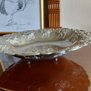 Op de afbeelding: Een zilverkleurige, ovaalvormige serveerschaal met sierlijke bloemen- en bladmotieven langs de rand. De schaal staat op kleine voetjes en weerspiegelt het oppervlak eronder. Op de achtergrond is een ingelijste schets te zien.