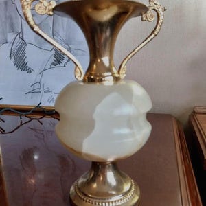 May include: A decorative vase with a gold-colored metal base and rim, and two ornate handles. The central body of the vase is a creamy white color. The vase has a bulbous shape and a wide opening.