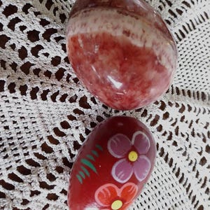 Pode incluir: Duas pedras lisas de formato oval. A pedra superior apresenta uma aparência polida e marmorizada com faixas em vermelho escuro, branco e castanho. A pedra inferior é de um vermelho escuro sólido, pintada à mão com duas flores rosa e laranja.
