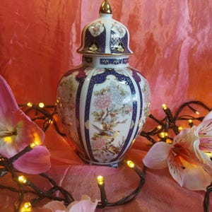 Op de afbeelding: Een decoratieve keramische vaas met deksel, met bloemen- en vogelmotieven in roze, blauw en goud. De vaas heeft een witte basis met ingewikkelde patronen en een gouden knop op het deksel. De achtergrond is een zachte roze stof.