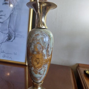 May include: An elegant vase with a brass top and base, featuring a marbled pattern in shades of brown, beige, and green. The vase has a unique shape and a decorative handle. A framed portrait is in the background.