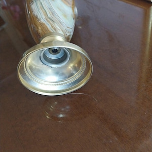 May include: A decorative vase with a gold-colored metal base and a patterned body in shades of brown, beige, and white. The vase has a wide, circular base and a narrower neck. The image is taken from a low angle, highlighting the base and the texture of the vase.