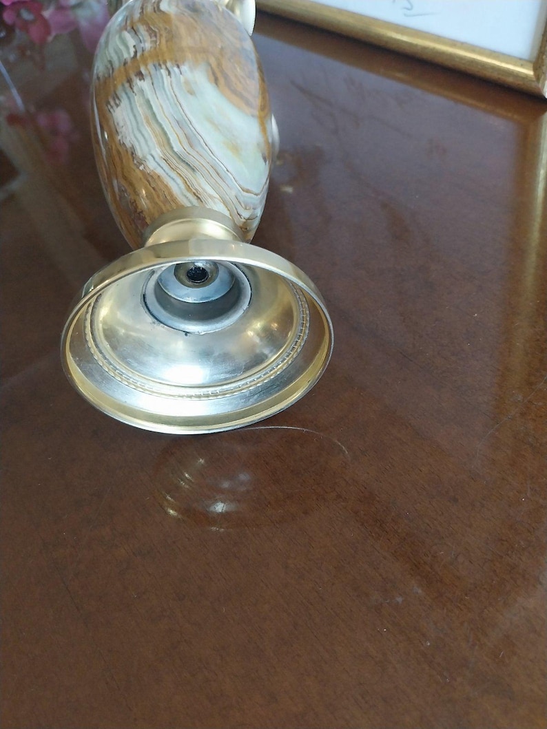 May include: A decorative vase with a gold-colored metal base and a patterned body in shades of brown, beige, and white. The vase has a wide, circular base and a narrower neck. The image is taken from a low angle, highlighting the base and the texture of the vase.