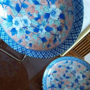 May include: Two round ceramic plates featuring a peach-colored background with a repeating pattern of blue and white flowers and green leaves. The edges of the plates are decorated with a dark blue and white geometric grid pattern border.