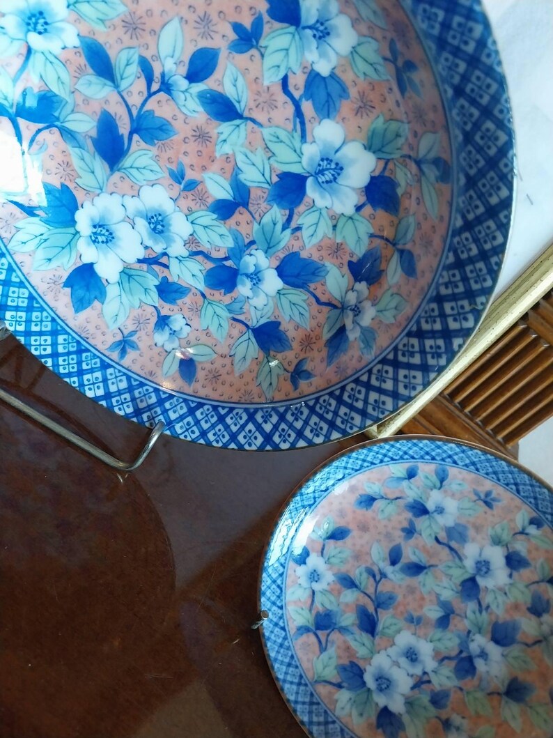 May include: Two round ceramic plates featuring a peach-colored background with a repeating pattern of blue and white flowers and green leaves. The edges of the plates are decorated with a dark blue and white geometric grid pattern border.