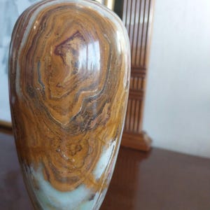 May include: A decorative vase with a polished, swirling pattern of brown, tan, and white tones. The vase has a brass-colored top and base. The overall shape is a rounded body that tapers towards the base.