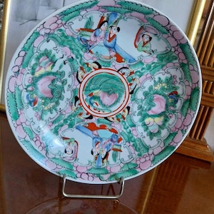 Peut inclure: Une assiette décorative en porcelaine avec un motif coloré et détaillé. L'assiette présente un médaillon central avec des motifs d'oiseaux et de fleurs, entouré de personnages en tenue traditionnelle et d'une bordure rose et verte.