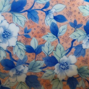 May include: A ceramic surface featuring a repeating pattern of blue and white flowers with green leaves set against a peach-colored background. The design includes small dark brown circular dots and star-like accents scattered throughout the floral arrangement.