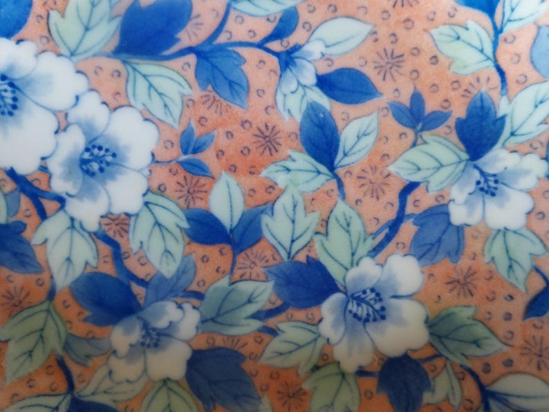 May include: A ceramic surface featuring a repeating pattern of blue and white flowers with green leaves set against a peach-colored background. The design includes small dark brown circular dots and star-like accents scattered throughout the floral arrangement.